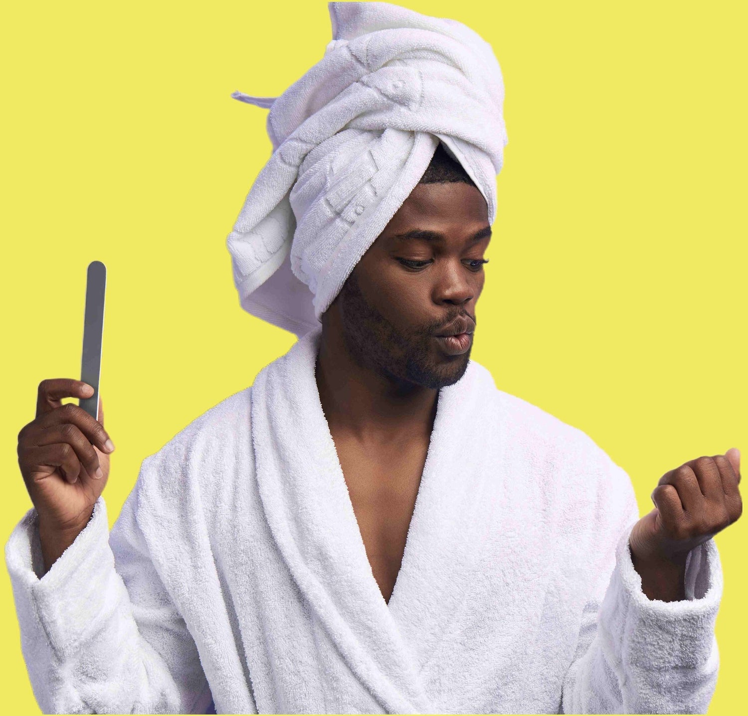 Black man with a white towel on his head and wearing white gown showing off his manicure at home, looking at his natural nails.